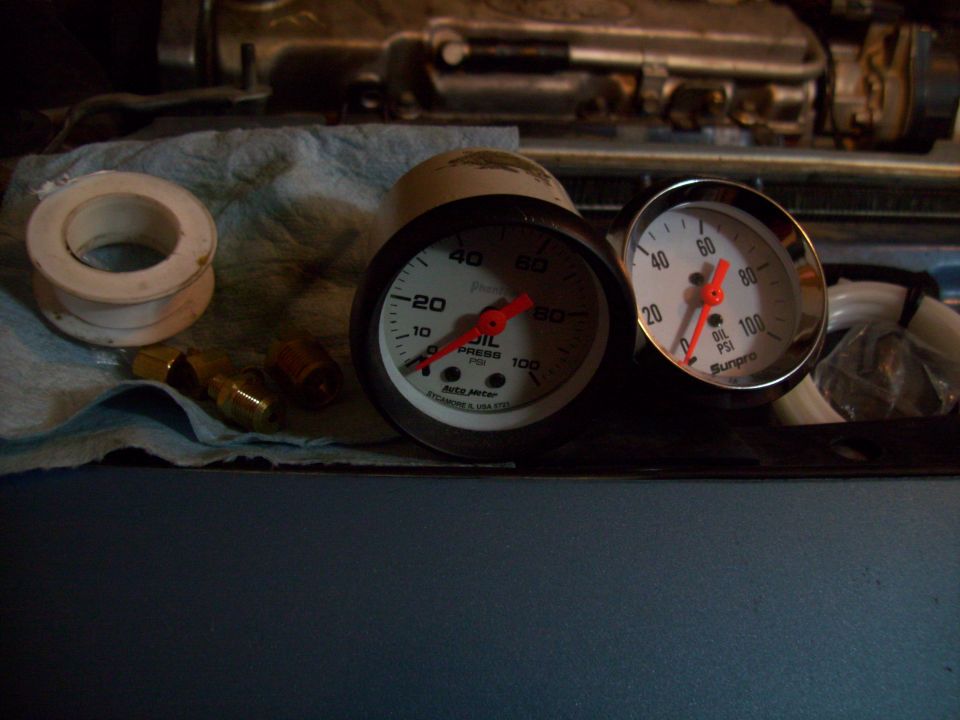Two oil pressure gauges and plumbing fittings on a blue surface with engine parts in the background