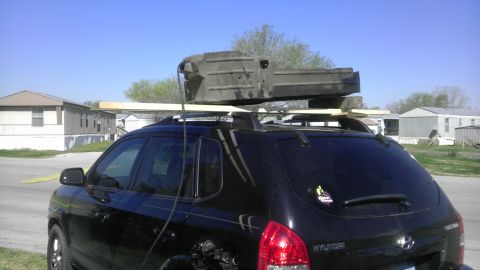 1982 Jeep Wagoneer - A black SUV is seen from the rear, carrying a large, dark object secured to its roof rack. The Supermotors.net logo is visible on the rear…