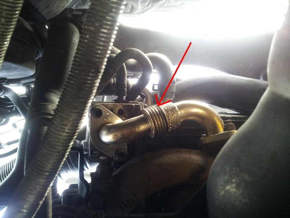 Close-up of a turbocharger pipe with a red arrow pointing to a corrugated section. Engine components and hoses are visible