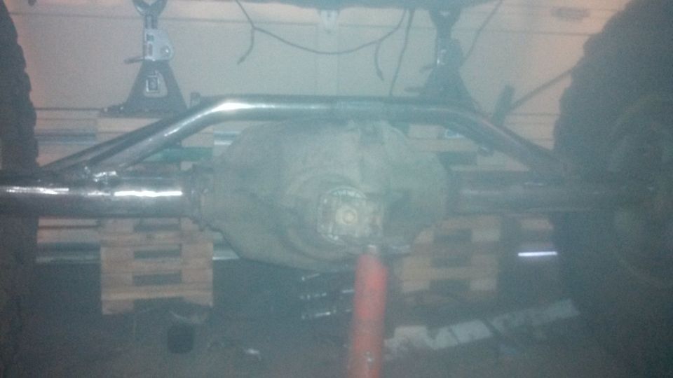 Rear axle assembly with suspension bars and tires visible, supported by jack stands and wooden pallets