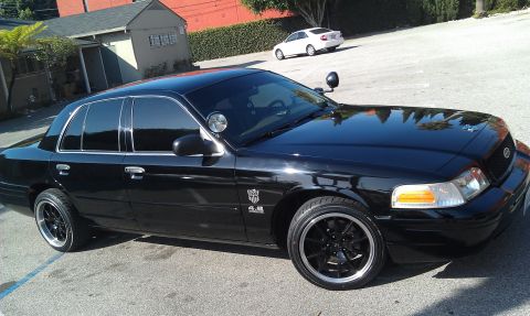 2003 Ford Crown Victoria - Murdered-out black full-size sedan, decklid emblem and black alloy wheels, front three-quarter view.