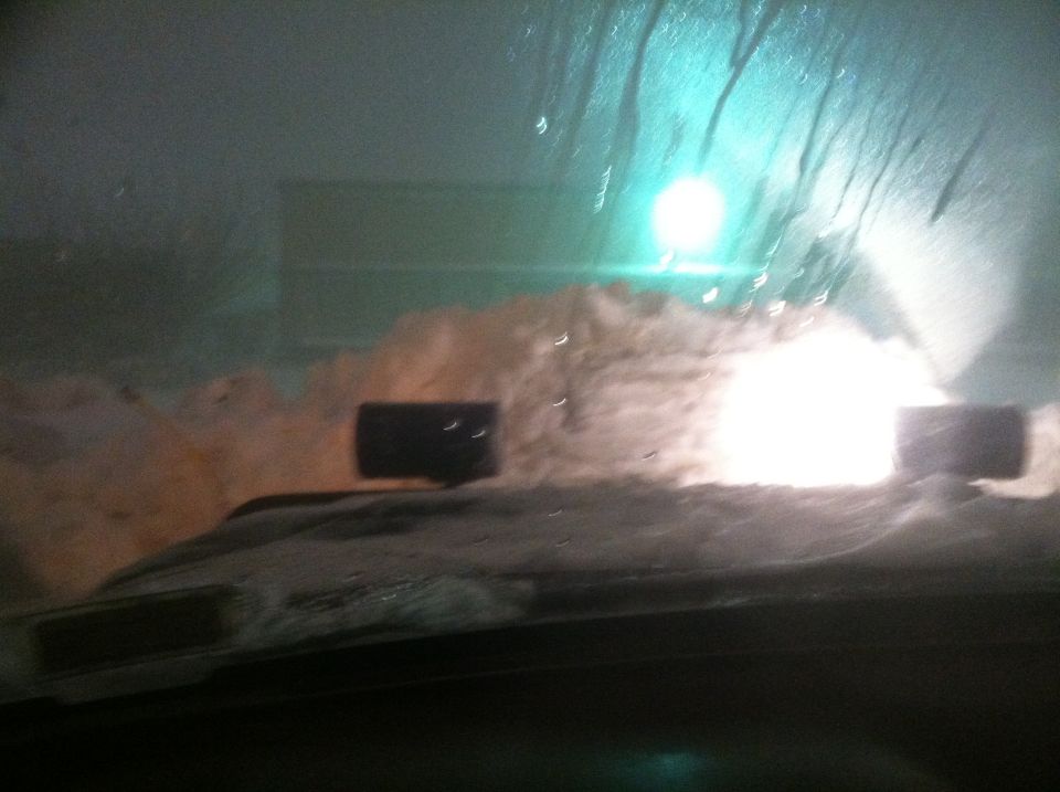 Dashboard view through a snow-covered windshield with bright lights ahead