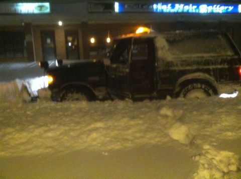 1989 Ford Bronco - A dark-colored Ford Bronco is stuck in a snowdrift at night, with its headlights on.