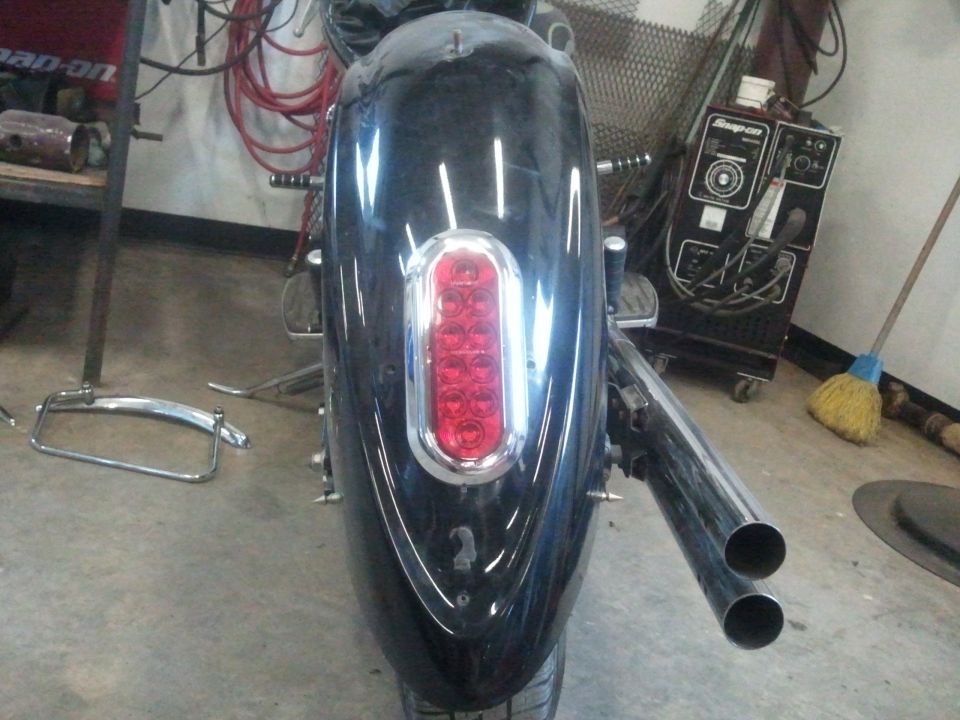Motorcycle rear fender with a chrome-trimmed LED taillight and dual exhaust pipes. A welding machine is visible in the background
