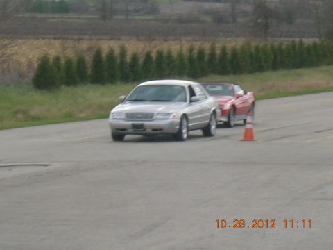 2003 Ford Crown Victoria - A silver 2003 Ford Crown Victoria sedan is followed by a red convertible on a paved track.