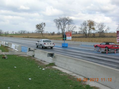 2003 Ford Crown Victoria - Two cars race down a drag strip under a cloudy sky, with a traffic light overhead.