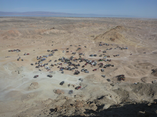 Many off-road vehicles parked in a desert valley under a clear blue sky