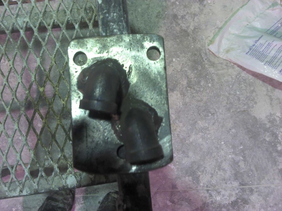 Metal plate with two black pipe fittings, one broken, on a concrete floor next to a metal grate