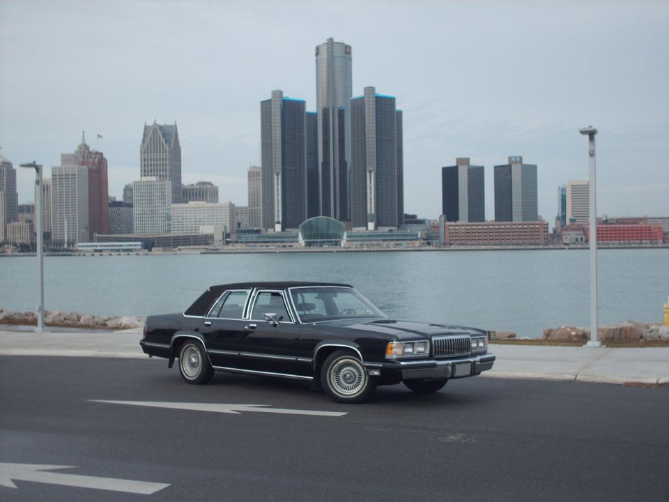 Black sedan parked by the water with a city skyline in the background