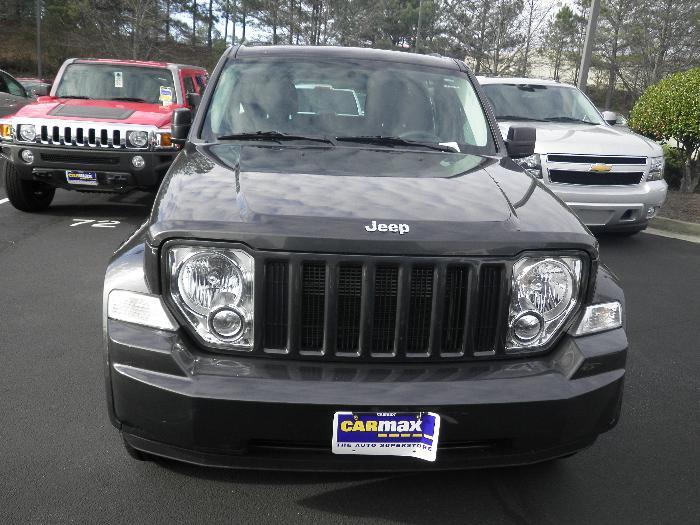 Front view of a dark gray Jeep Liberty with a CarMax license plate frame. A red Hummer and a silver Chevrolet Tahoe are parked behind it