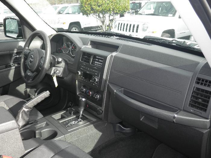 2011 Jeep Liberty interior showing dashboard, steering wheel, center console, and gear shifter. Two white SUVs are visible through the windshield