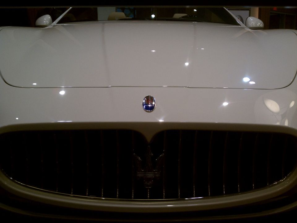 White Maserati front grille with trident emblem and oval badge above. Supermotors.net watermark in lower right