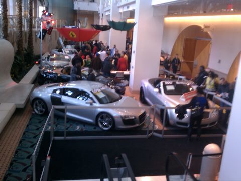 1998 Toyota Camry - Two luxury sports cars are showcased at an indoor event, with spectators milling around.