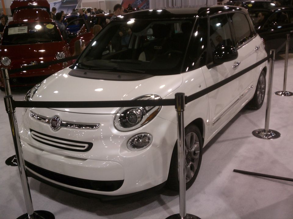 White Fiat 500L with black roof and trim, parked indoors with stanchions. Red Fiat 500 visible behind it