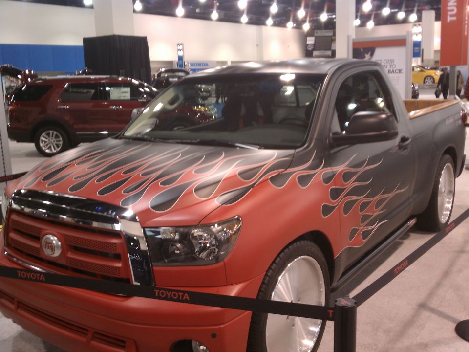 Toyota Tundra pickup truck with red and black flame graphics on the hood and sides. It has a custom grille and large chrome wheels. Another SUV is visible in the background