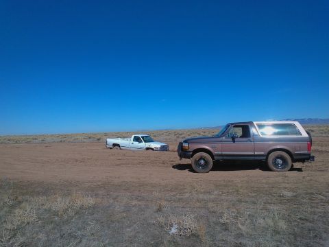 1992 Ford Bronco - A brown and white 1992 Ford Bronco sits on dry ground while a white pickup truck is stuck in the mud in the distance.