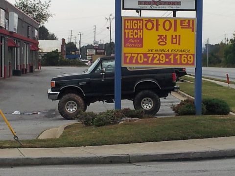 1986 Ford Bronco - A black pickup truck with large tires is parked next to a business with a multilingual sign.