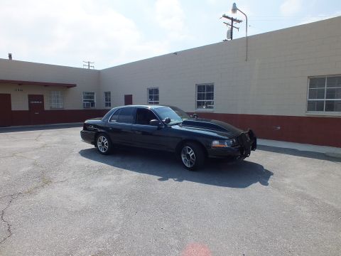 2003 Ford Crown Victoria - Black full-size sedan with tinted windows on silver wheels, parked beside a warehouse wall.