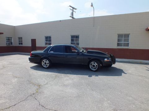 2003 Ford Crown Victoria - Side profile of a black full-size sedan on silver alloy wheels against a tan masonry building.