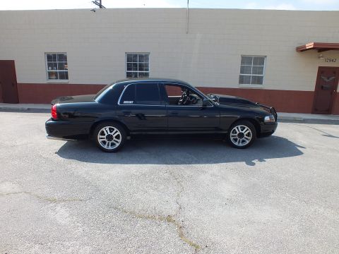 2003 Ford Crown Victoria - Black full-size sedan in profile, slightly lowered stance, parked outside an industrial wall.