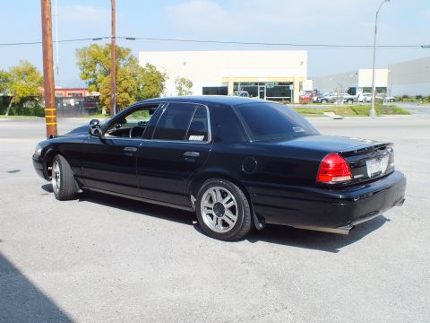 2003 Ford Crown Victoria - Black full-size sedan from the left rear quarter on silver wheels in a commercial lot.