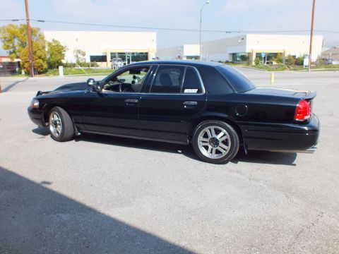 2003 Ford Crown Victoria - Driver-side rear quarter of a black full-size sedan parked outside an industrial building.
