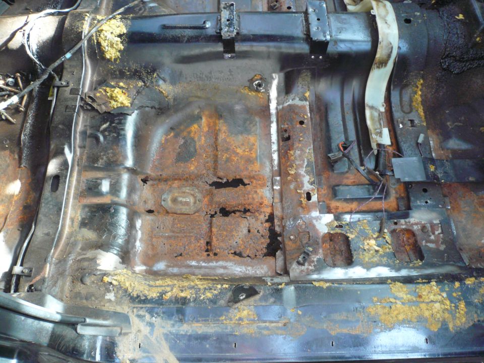 Heavily rusted car floor pan with holes and yellow insulation visible