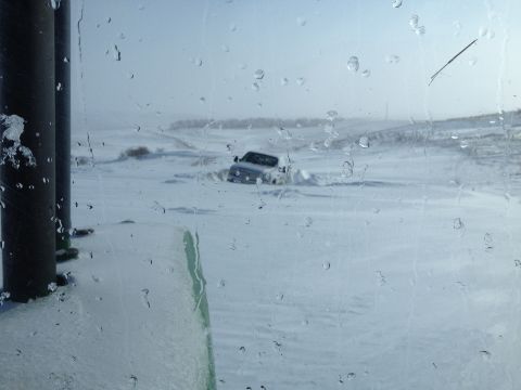 1979 Ford Bronco - A white SUV is stuck in a snowdrift, viewed from inside a vehicle through a window covered in raindrops.