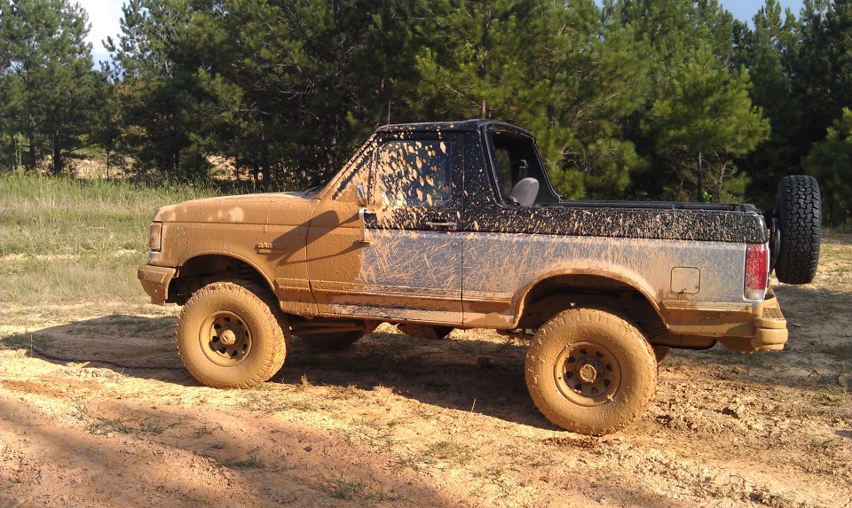 Mud-covered 1990 Ford Bronco parked on dirt with trees in background
