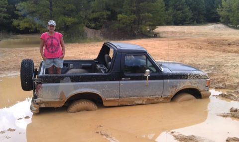 1990 Ford Bronco - A woman stands in the bed of a two-tone Ford Bronco that is stuck in a muddy area.