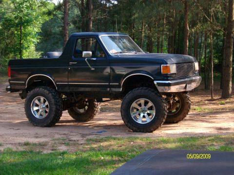 1989 Ford Bronco - This lifted Ford Bronco is parked in a wooded area. The vehicle has large off-road tires and chrome wheels.