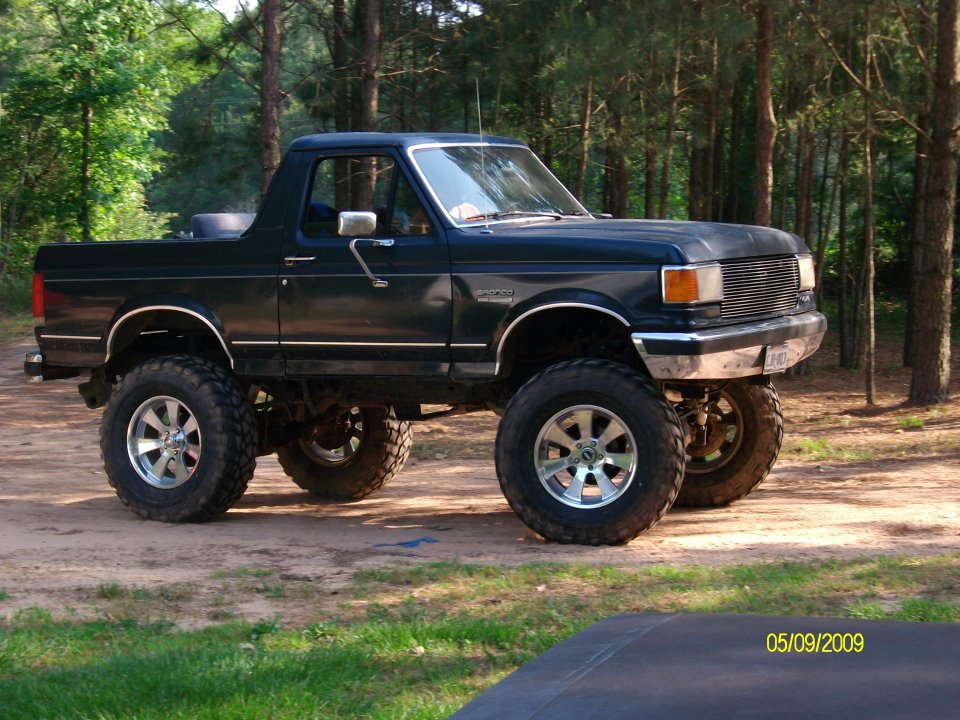 Lifted black Ford Bronco parked on dirt road with trees in background. Has aftermarket wheels and tires