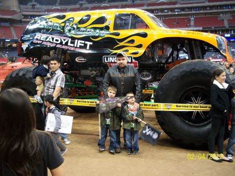 1989 Ford Bronco - A monster truck with a flame graphic and "Ready Lift" branding is shown with families posing in front of it.