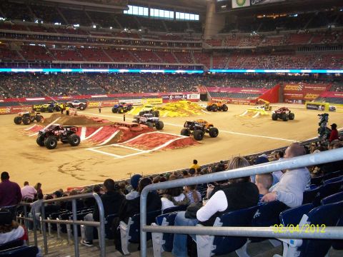 1989 Ford Bronco - Monster trucks are prepared for an event in a stadium, with ramps and obstacles on the dirt arena.