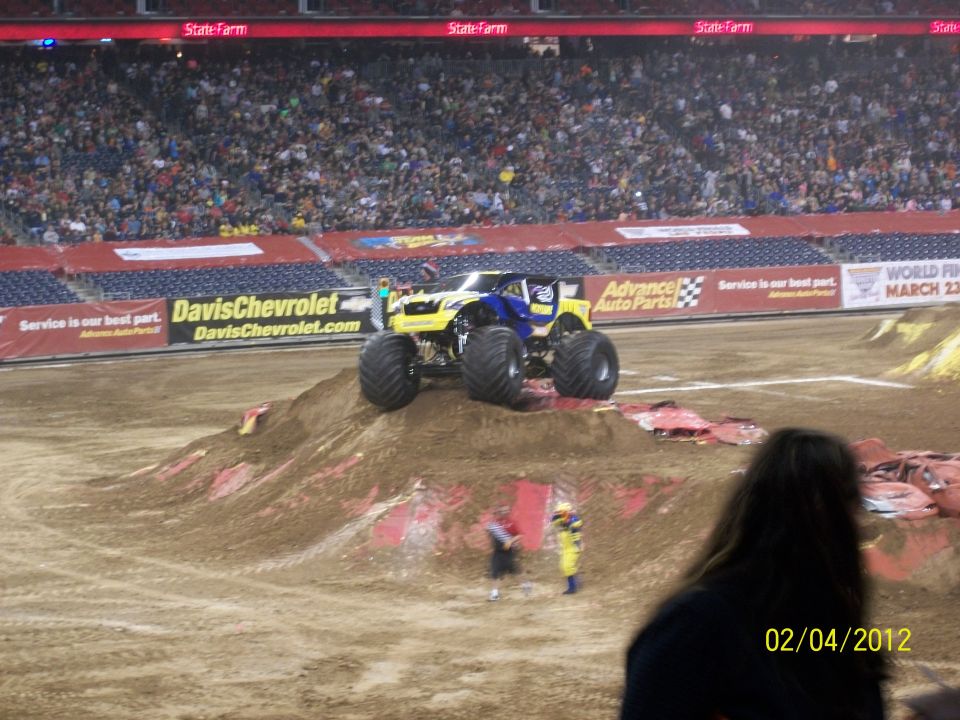 Monster truck with yellow and blue body on dirt ramp, large tires, stadium seating in background