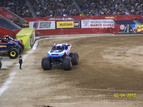 1989 Ford Bronco - A monster truck with a patriotic stars and stripes design sits on a dirt arena floor, with other vehicles and spectators in the background.