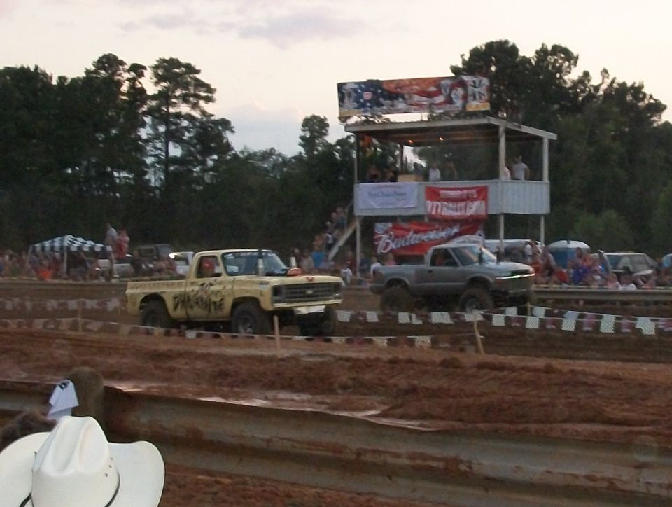 Two trucks racing in a mud pit with spectators and a grandstand in the background