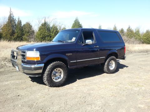 1995 Ford Bronco - A dark blue Ford Bronco is parked on a dirt road under a clear sky.