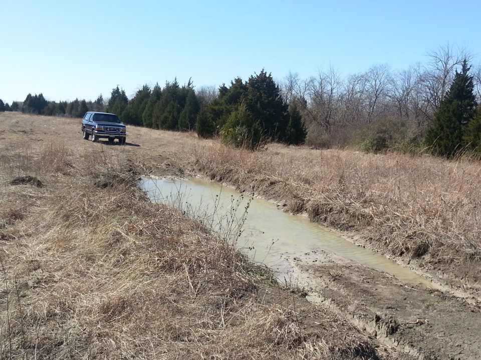 Blue Ford Bronco driving on a dirt path next to a muddy ditch
