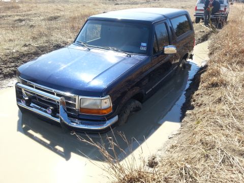 1995 Ford Bronco - A blue SUV is stuck in a muddy rut, with another vehicle and people nearby.