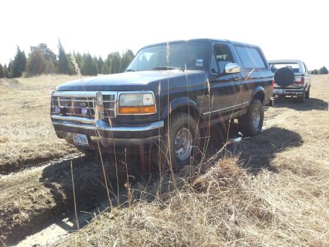 1995 Ford Bronco - A dark blue Ford Bronco is stuck in a muddy field with another truck visible in the background.