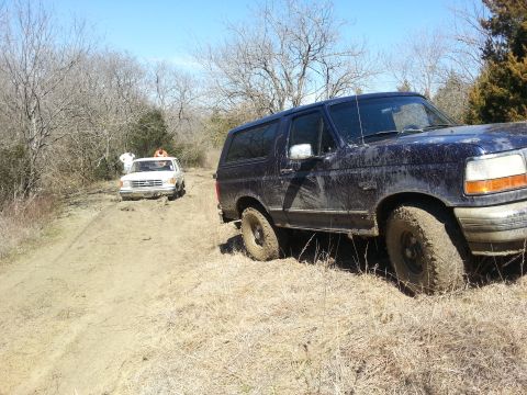 1995 Ford Bronco - Two SUVs are stuck in a muddy, wooded area on a sunny day.