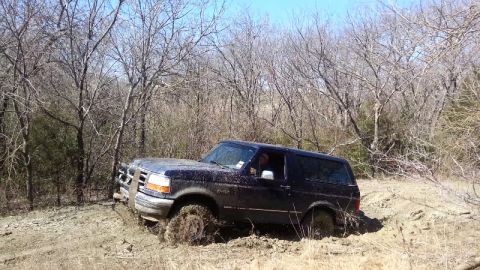 1995 Ford Bronco - A Ford Bronco navigates a muddy off-road path surrounded by bare trees under a clear sky.