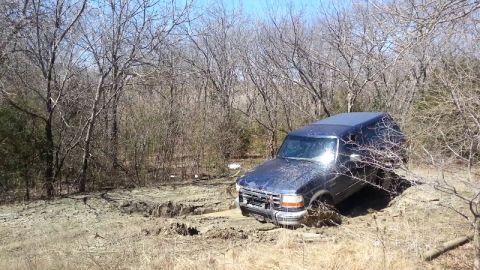 1995 Ford Bronco - A blue 1995 Ford Bronco is stuck in the mud off-road, surrounded by bare trees under a clear sky.