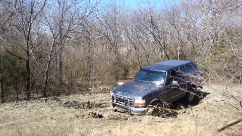 1995 Ford Bronco - A dark blue 1995 Ford Bronco is stuck in the mud, surrounded by trees and dry brush.