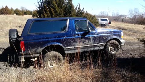 1995 Ford Bronco - A blue Ford Bronco navigates a muddy off-road trail, with another vehicle visible in the distance.
