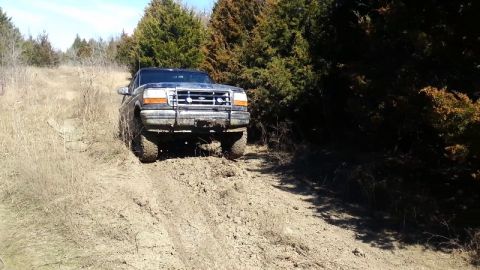 1995 Ford Bronco - A Ford Bronco navigates a muddy trail through a wooded area on a sunny day.
