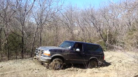 1995 Ford Bronco - A dark blue Ford Bronco is stuck in thick mud off-road, surrounded by bare trees.