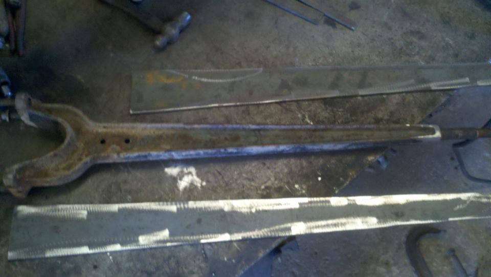 Rusty suspension component with two mounting holes, lying on a workshop floor with metal plates