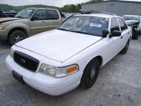 2003 Ford Crown Victoria - White 2003 Ford Crown Victoria police car in a junkyard with a tan Ford Explorer nearby.